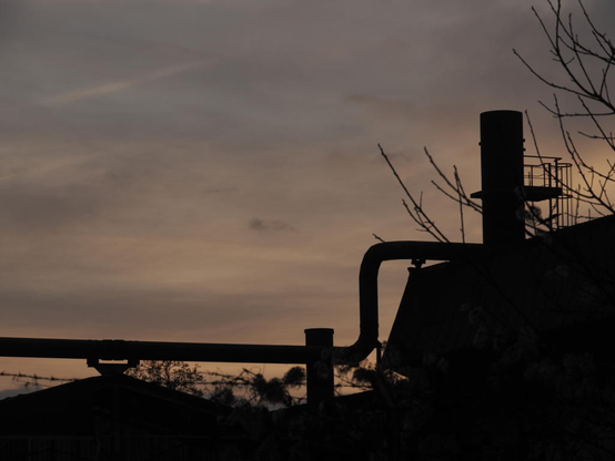 Vor einem Himmel mit Wolken die noch gelborgange von der Sonne angestrahlt sind sieht man Dächer und Rohre der Hüttengebäude als Shilouette.