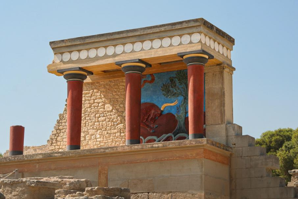 A partial view of a Minoan temple reconstruction featuring red columns, a colorful fresco of a bull, and stone walls under a clear blue sky.