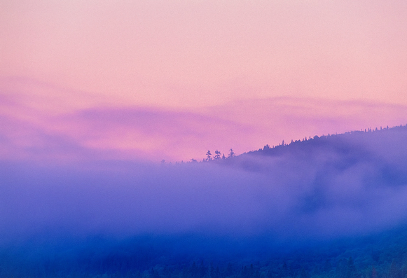 Low cloud mixes with fog rising from a lake surrounded by hills under a pink and purple sunrise sky. 