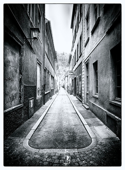 Kleine Gasse neben einer Einkaufsstrasse ohne deren Glanz.  Triste Fassaden, zugemauerte Fenster. Im Verlauf rechts ein Werbeschild über einer Tür.