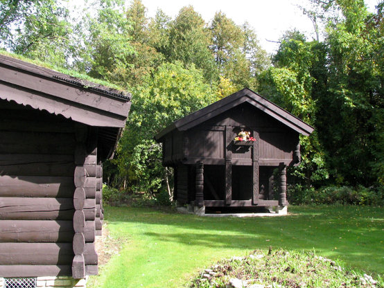 Two log cabin sod-roofed buildings on the grounds of Al Johnson's in Sister Bay, a number of years ago, with green grass and trees.