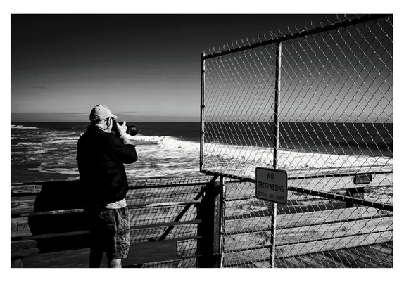 A person in casual attire stands on a pier, photographing the ocean waves with a camera. A "No Trespassing" sign hangs on a chain-link fence nearby.