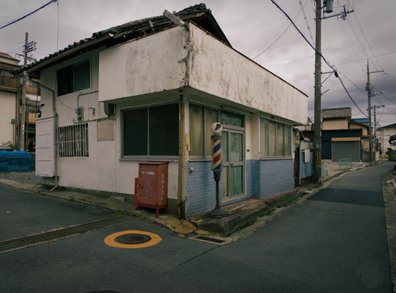 This image shows a small, two-story building at a street intersection. The building has a white exterior with blue tile on its ground floor and a sloped roof. A red-and-blue barber pole stands near the entrance, which features a glass door and several windows. A red metal mailbox is positioned adjacent to the pole. The building occupies the corner where two paved streets meet; one street has a circular yellow marking around the manhole. Utility poles with overhead wires are visible in the background, and other residential structures are present under an overcast sky. The building shows signs of aging, with visible wear on the exterior.