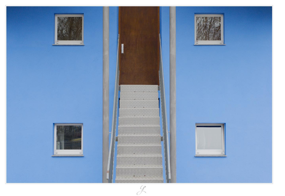 The facade of a blue house, right upfront. It is quite symmetrical: in the center top, there is a silver-gray metal stair leading straight to a  dark brown wooden door at the upper end. Left and right to the steps there are silver-gray metal railings. At the bottom of the stairs, there are two thin gray metal pillars at the end of the railings reaching from the ground straight up to the roof, one to the left and the other to the right. Left and right of the pillars, four small symmetrically ordered squared windows can be seen, each of it exactly marking the four corners of a larger square reaching from the ground floor to the first floor (perhaps not exactly a square, but it seem as if it could be a square).

AI disclaimer: Using my work, its meta data, written or derived description to create media with or train AI based systems is prohibited.