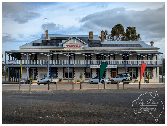 A wide shot of the two story Dumbleyung Tavern, a historic stone building with a corrugated iron roof covered in solar panels.  A wrought iron verandah runs along the front of both floors. Several vehicles, including a white sedan and a white SUV, are parked in front of the ground floor.  The gravel and asphalt parking area is in the foreground, with three advertising flags (green, white, and red) marking the curb. The sky is partly cloudy.