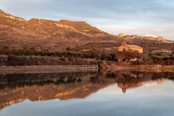 Español: paisaje rural al atardecer en Arascués, Huesca. Frente a un lago tranquilo se refleja la Ermita de la Virgen del Olivar, rodeada de árboles y vegetación. Al fondo se ven colinas y montañas iluminadas por la luz dorada del sol.

English: rural landscape at sunset in Arascués, Huesca. The calm lake reflects the Hermitage of the Virgin of Olivar, surrounded by trees and natural vegetation. In the background, hills and mountains are lit by warm golden light.