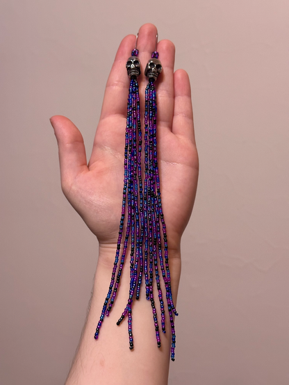 My hand holdng up a pair of long beaded dangle earrings in sparkly blue, purple, and black, with silver skull beads at the top