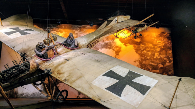 A dynamic museum display at the Omaka Aviation Heritage Centre in Blenheim. Two full-scale, historical replica World War I German aircraft (recognizable by the black Iron Cross markings on the wings) are suspended, depicting an aerial dogfight over a battlefield.

In the foreground, a large, two-seater biplane (likely a German Aviatik) is angled sharply, showing the pilot and the gunner (who is operating a machine gun from the rear cockpit). The biplane has a beige, worn canvas finish and detailed engine components visible.

In the background, a smaller biplane (a German fighter like a Fokker or Albatros) is flying further away, silhouetted against a dramatic, back-lit wall mural depicting an orange-and-yellow sky suggesting fire and smoke over an aerial photo of trenches and fields below.