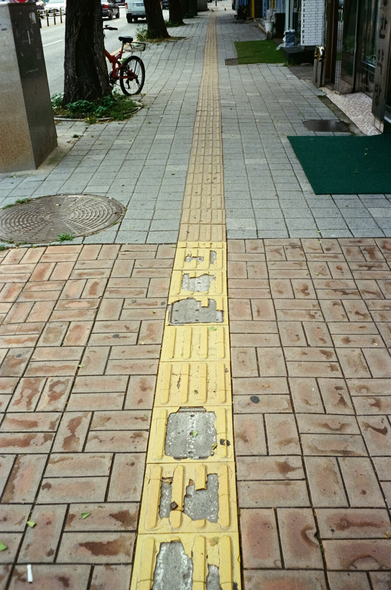 도심 인도바닥에 노란색 점자블록이 일자를 그리며 설치되어있다. 보도블럭이 회색 영역과 갈색 영역으로 구분되어 있는데, 점자블록도 오래되어 낡고 색이 바랜 부분과 너무 낡아서 망가진 부분이 선명하게 구분된다.