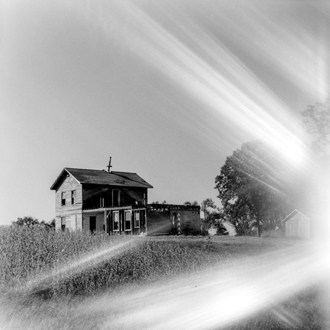 A black and white photo of a dilapidated farm house standing in a field of long grasses. The farm house is two stories with an extension on the side that is a single story which somewhere between unfinished and disrepair. There's a large tree in the background alongside a shed. The entire photo is very heavily affected by light leaks emanating from the middle right and streaking across the frame both up and down in a V shape almost as if destroyed the house and now is sweeping across the landscape.