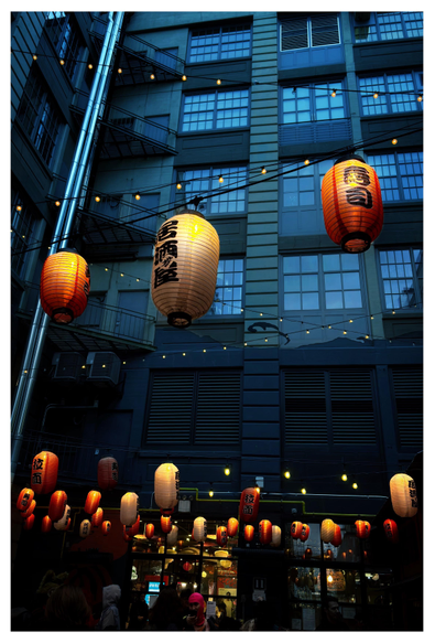 Photo of the courtyard of Japan Village in the Industry City complex in Brooklyn, looking up at the facade of a high-rise building. Capsule-shaped illuminated lanterns with kanji characters, in alternating colors of white and red-orange, are suspended over the courtyard among crisscrossing wires carrying smaller yellow lightbulbs. People mill around in front of the glass doors at bottom frame. Contrasting with the warm glow of the lanterns are the cool blue tones of the facade, with windows reflecting the early evening sky.