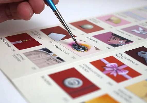 A hand holds a fine brush over a color palette, painting over the "Pink Ground" shade on a paint sample color chart.