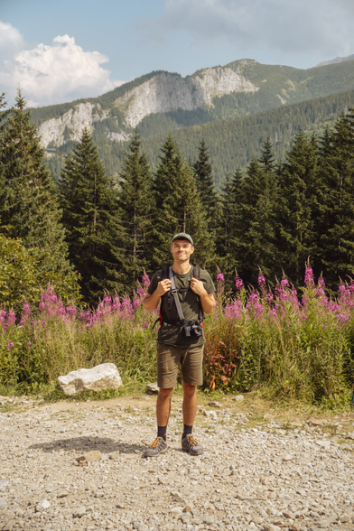 A photo of me with a black Yashica analog camera, with a picturesque view of mountains, coniferous forest, and pink fireweed flowers filling the frame in the background. The sky is cloudy. Wooden houses stand along the path.
