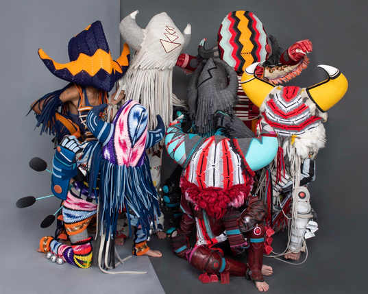 a series of costumed dancers wearing sculptural pieces by Cannupa Hanska Luger, assembled in a group against a grat background