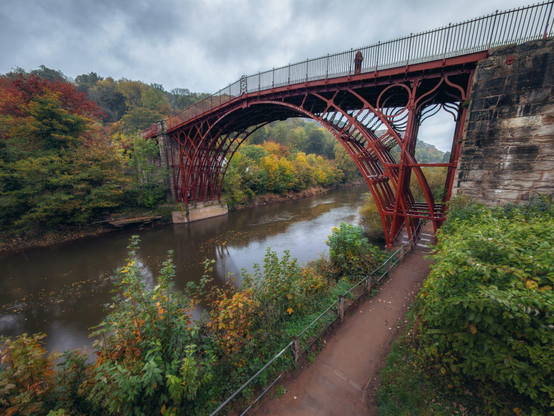 A view of the Iron Bridge shows its red cast iron ribs arching gracefully over the River Severn, framed by dense autumn foliage in shades of green, gold and deep red. The river runs quietly beneath the structure, reflecting patches of sky and scattered leaves. A narrow riverside path curves below the right side of the bridge, bordered by shrubs and a metal handrail. A person stands on the bridge, small against the ornate latticework and stone abutments. Low cloud and soft light give the scene a calm and misty atmosphere.