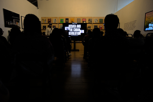 Photo taken from inside the commemoration. At a low angle through the crowd is visible a screen with the words "Assata and the Cuban Revolution".