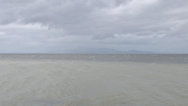 A photo of a large fjord with mountains in the distance. The water distinctly has two different colors to it, with paler more yellow toned water closer to the camera. The sky is filled with clouds.