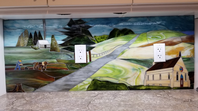 First of three panels of stained glass depicting the view out my window as it would have been in the past. In the foreground to the left is a farmer steering and pushing an old single blade plough being pulled by a sturdy horse. In the background is his log cabin, nestled among pine trees, with smoke coming out of the chimney. An angled grey road with a fence line running beside it divides the image from a country church in the foreground to the right. Fields in the green colours of spring lay behind it.