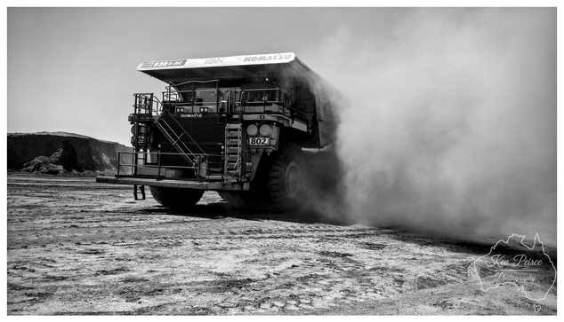 A dynamic, low angle, black and white photograph signed by Kev Peirce. A massive Komatsu 830E-AC haul truck, identified by its model number and '802' on the side, is seen from the rear quarter.  The truck is driving on a dusty, cracked dirt surface, generating a huge, thick cloud of dust that billows dramatically to the right side of the frame.  In the background, there is a large, sheer embankment of excavated material. The image captures the scale and activity of a major mining operation.