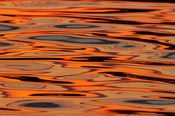 Photograph of sunset colors of bright orange, blue, and black reflected on water forming horizontal swirls and patterns.