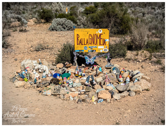 A wide angle photo of a yellow wooden sign in a dry, scrubby roadside environment. The sign, hanging between two posts wrapped in fairy lights, reads "BALLAGNOMEIA" in large orange, whimsical text.

Below the sign is a large cairn or pile of rocks covered in dozens of small garden gnomes and various trinkets, including a blue butterfly decoration, a small toy goat, and some hats. The ground is dry dirt and gravel.
