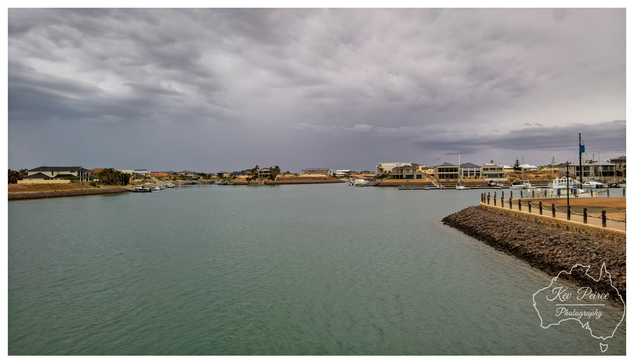 A wide angle landscape photograph of the Wallaroo Marina. The scene features dark, turquoise coloured water under a heavy, overcast, and moody grey sky.  Waterfront homes line both sides of the marina channel. On the right, a rocky retaining wall and a fenced promenade lead into the distance where several boats are moored.