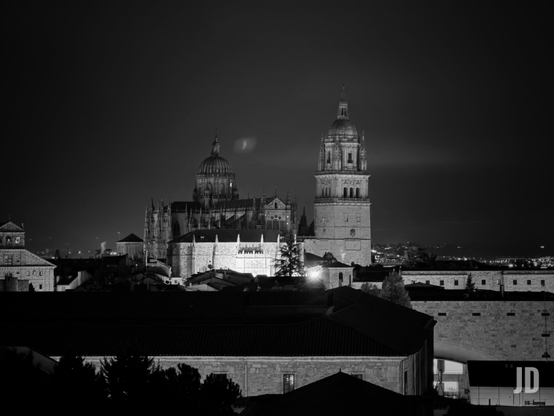 Fotografía nocturna en blanco y negro de un paisaje urbano. En el centro, una imponente catedral o complejo religioso iluminado destaca contra el cielo oscuro. Presenta una gran cúpula prominente a la izquierda y una alta torre campanario con cúpula más pequeña a la derecha, ambas con arquitectura elaborada. Por debajo, se ven los tejados y fachadas parcialmente iluminadas de otros edificios de la ciudad en primer plano, creando varias capas de siluetas oscuras. En el horizonte, se aprecian las luces tenues de la ciudad extendiéndose a lo lejos. El contraste entre la luz de los edificios y la oscuridad del cielo y los primeros planos domina la imagen.