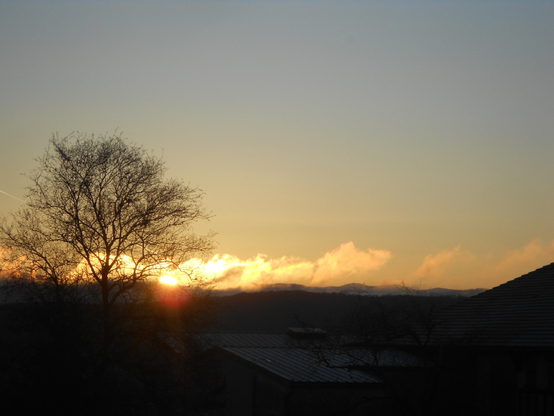 L'horizon montagneux Est où le soleil parait déjà à moitié parmi quelques nuages blanc. Au premier plan la silhouette à contre-jour d'un arbre dénué de feuilles. 