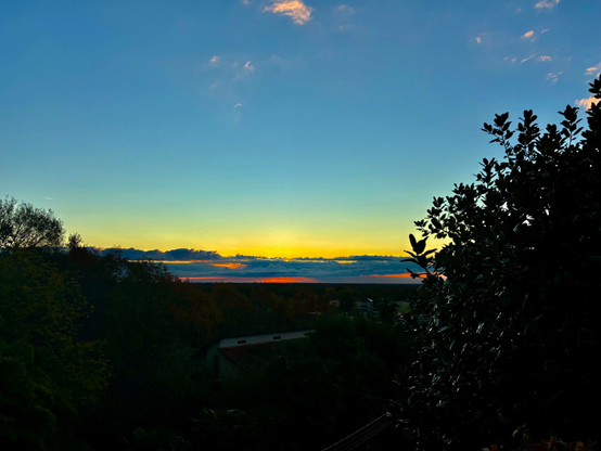 A serene sunrise  view with a gradient sky transitioning from blue to warm hues of orange and yellow. Silhouetted trees are visible in the foreground, enhancing the tranquil scene.