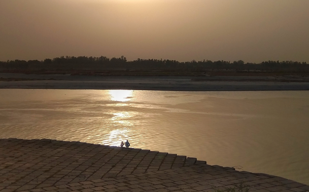 Sunset over a calm river, with golden reflections on the water. A silhouetted man with child sits on cobblestones.