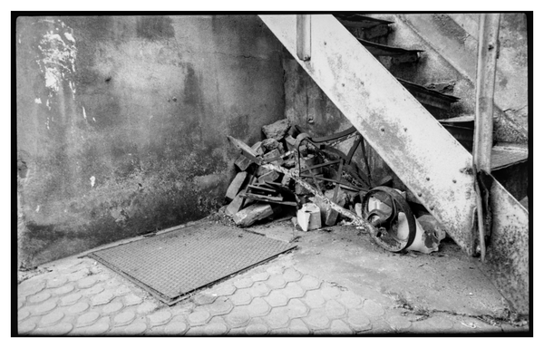 Black and white photograph in landscape orientation. The remnants of a broken wheelbarrow sit on top of some stones under an old metal stair.