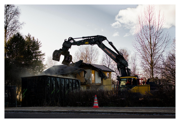 En liggande bild på ett gult arbetsfordon med grävskopa som används för att riva ett gult trähus. Det ryker ur en mörk container och på trottoaren framför står en randig orange kon. Träd runtom och en blå himmel med ett vitt moln uppe till höger.