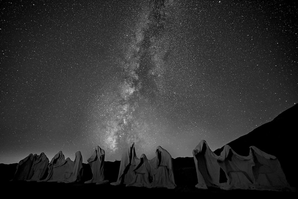 A black and white landscape photo of a line of statues depicting Da Vinci’s painting, The Last Supper, only these figures are white ghosts. The photo was made under the night sky with the Milky Way shooting up from through the center of the photo. 