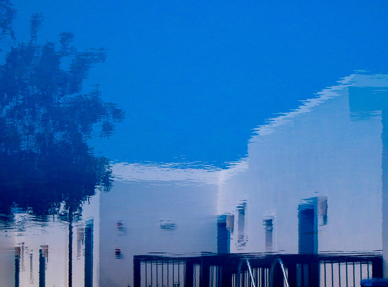 An abstract reflection of a building and trees on a water surface, creating a distorted image against a blue sky backdrop.