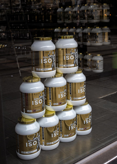 Photo of a storefront selling fitness food supplements. There is a pyramid-shaped stack of massive jars of protein powder with shiny golden lids and labels reading "GOLD ISO". Behind it, there are more and more shelves of the same thing fading out into the distance. In the reflection on the glass of the storefront, a ladder is faintly visible, leading up towards the top of the frame.