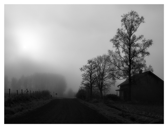 En liggande kontrastrik svartvit bild på en grusväg med ett hus till höger. Bakom huset står några träd. Till vänster om vägen ett staket och in i dimman syns ett trädridå.