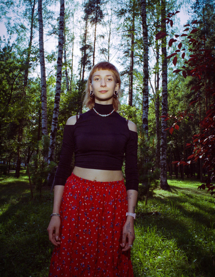 Color flm photo of a non-binary person with a red long skirt and black shirt and a sparse forest behind them, with somewhat noticeable usage of flash - the center of the image and the face is visibly lit
