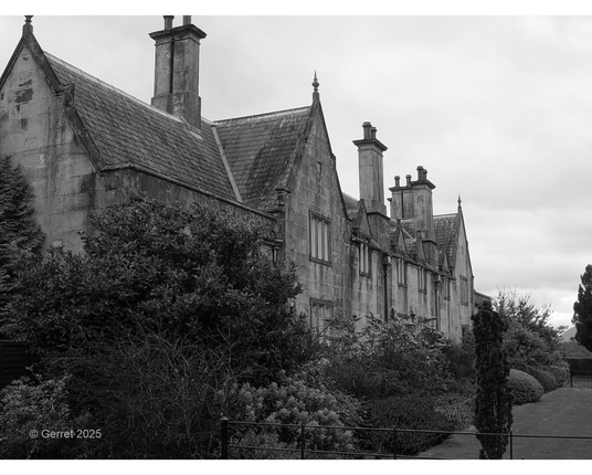 Historic stone mansion with tall chimneys, pointed gables, and lush gardens, captured in black and white, evoking a mysterious, serene atmosphere.