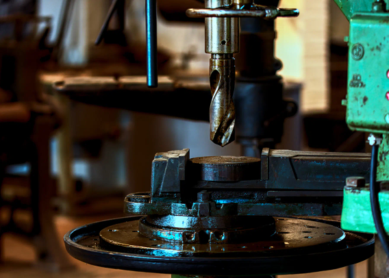 Ein großer, mindestens daumendicker Bohrer hängt von oben herab. Unter ihm befindet sich eine Anordnung, um das Werkstück auf dem Gerät zu befestigen. Rechts ist ein wuchtiger Teil der Borhmaschine zu sehen, an dem Teller und Bohrer befestigt sind. 
Im Hintergrund sind eine Werkbank und vielleicht ein Stuhl zu erkennen. 