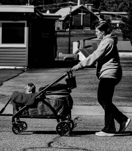 A black and white photo of a mother pushing a buggy on a city street.  The child in the buggy is peeking over the side looking at me snap the photo.