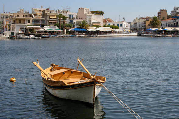 A small wooden boat is docked in a calm harbor with buildings and palm trees in the background. The scene features clear blue skies and water.