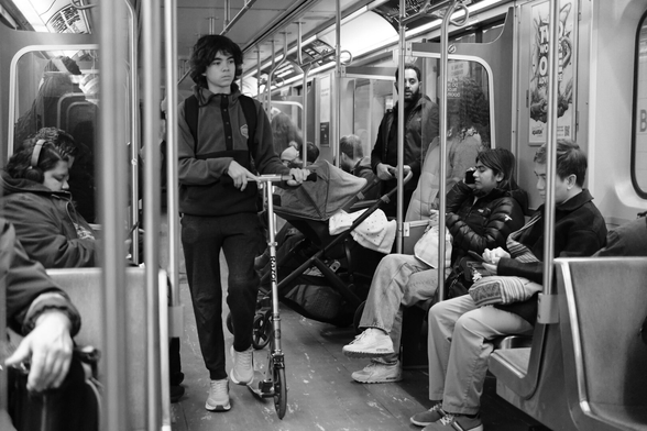 A crowded subway car. The seats are full. Standing by one of the doors is a young couple with a stroller. Twining between them all are the handrails and poles of the car’s interior.