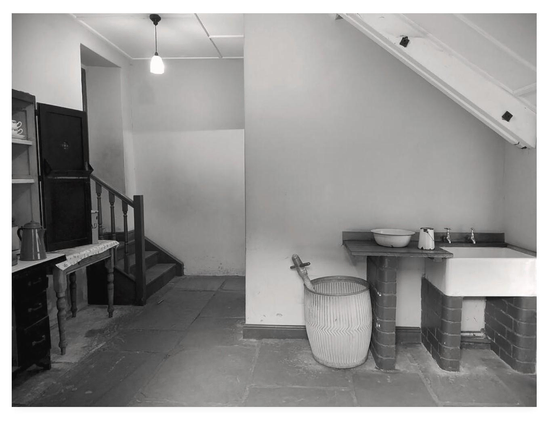 Black and white photo of an Edwardian scullery.  To the left, a crockery cabinet and coffee pot.  To the right, a Belfast sink.  Alongside the sink is a dolly tub and posser.  The floor is flagstoned.