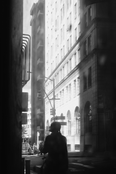 A silhouette is seen through the reflection of a dirty window. The silhouette is looking up and to the left. Beyond them is a brightly lit old stone building, street lamp, traffic signs, a car on the street.