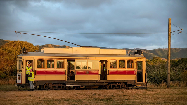 A side view of a vintage tram, labeled "180," at the beach end of a grassy track in a sunset rural setting. The tram is cream and deep red, with wooden window frames, and is a preserved historic electric trolley. Two people wearing high-visibility vests are standing on the left side near the open front entrance. In the background, there are low hills covered in vegetation, and the sky is overcast. A wooden utility pole with overhead wires is visible on the right.