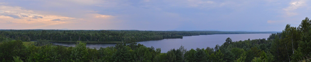 A panoramic photo of a large lake surrounded by forest. The sky is filled with purple clouds with some rays of light shining through.