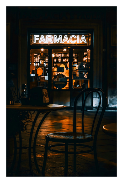 A dimly lit, cozy looking pharmacy at night. The neon sign "FARMACIA" glows brightly above the glass door. Inside, warm orange lights illuminate shelves of bottles and a counter. In the foreground, a black, wickered chair with a curved backrest sits at an empty, round wooden table. The glow of the lights are reflected on the damp street cobbles.