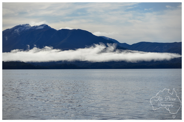 A dramatic landscape photograph taken across Macquarie Harbour. The foreground is the dark, textured surface of the water.

In the middle ground, a thick horizontal layer of white fog or low cloud hangs over the dark forested coastline.

The background is dominated by large, deep blue mountains with patches of snow or light cloud near their peaks, set against a pale blue and white sky. Photo by Kev Peirce.