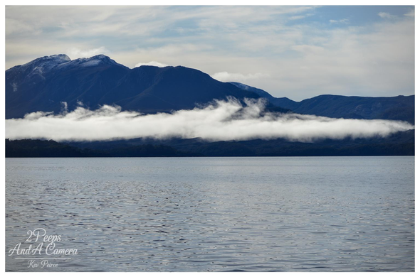 A dramatic landscape photograph taken across Macquarie Harbour. The foreground is the dark, textured surface of the water.

In the middle ground, a thick horizontal layer of white fog or low cloud hangs over the dark forested coastline.

The background is dominated by large, deep blue mountains with patches of snow or light cloud near their peaks, set against a pale blue and white sky. Photo by Kev Peirce.