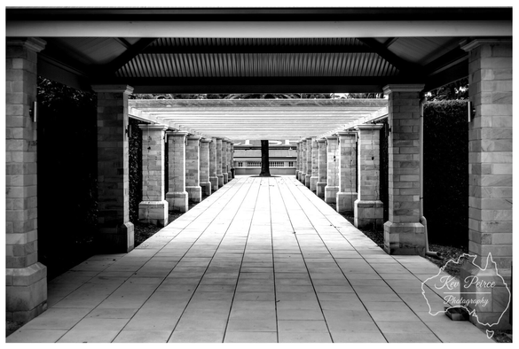 A high contrast black and white photograph of the long, colonnaded walkway at Seppeltsfield winery. The image uses strong central perspective, with two parallel rows of stone pillars receding sharply toward a single tree in the distance, framed by a building facade.  The tiled path leading down the centre emphasizes the depth and symmetry. The structure above features horizontal slats and a corrugated roof section at the near end. The photograph is signed 'Kev Peirce Photography.'
